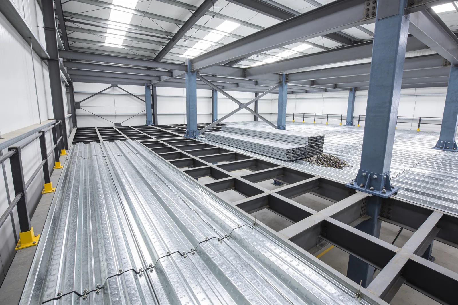 Industrial steel mezzanine under construction with corrugated galvanized B-deck panels laid and fastened over part of the joist grid, the remaining joists and beams still exposed