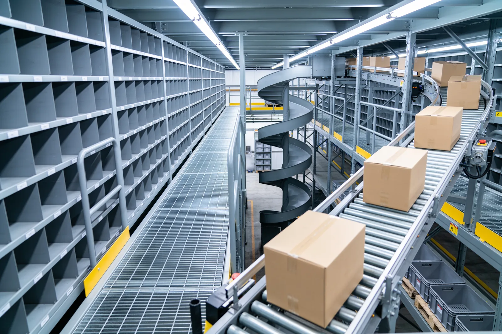 Elevated picking level with a bar-grating walkway between a shelving pick face and a roller take-away conveyor carrying cartons, with a spiral conveyor descending to the ground level in the background