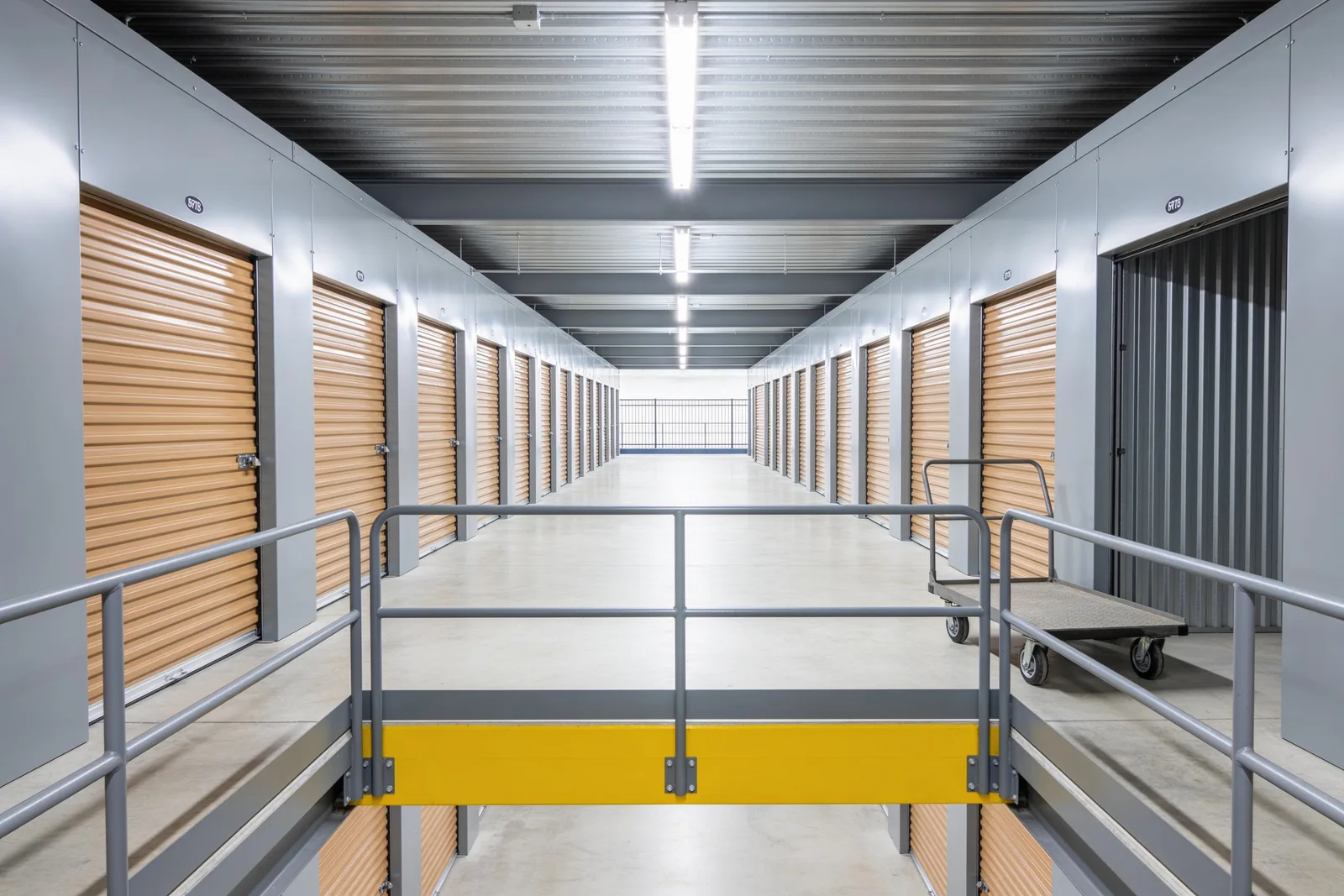 Upper level of a self-storage facility: a wide corridor of tan roll-up unit doors runs to the far wall under linear ceiling lighting, a grey steel guardrail with a safety-yellow kick plate protects the floor opening in the foreground, and a flatbed moving cart is parked beside one closed unit