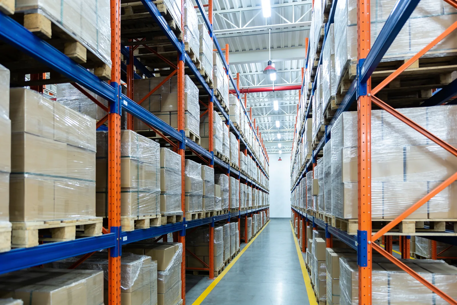 Narrow aisle running between tall selective pallet racking with orange beams and blue uprights, both sides fully loaded with shrink-wrapped palletised cartons stacked toward the roof structure