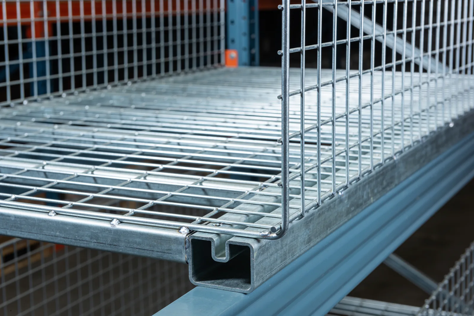 Close-up of a flat galvanized welded wire rack deck resting on a grey rack beam, its front edge folding down over the beam face with a formed steel support channel visible in cross-section