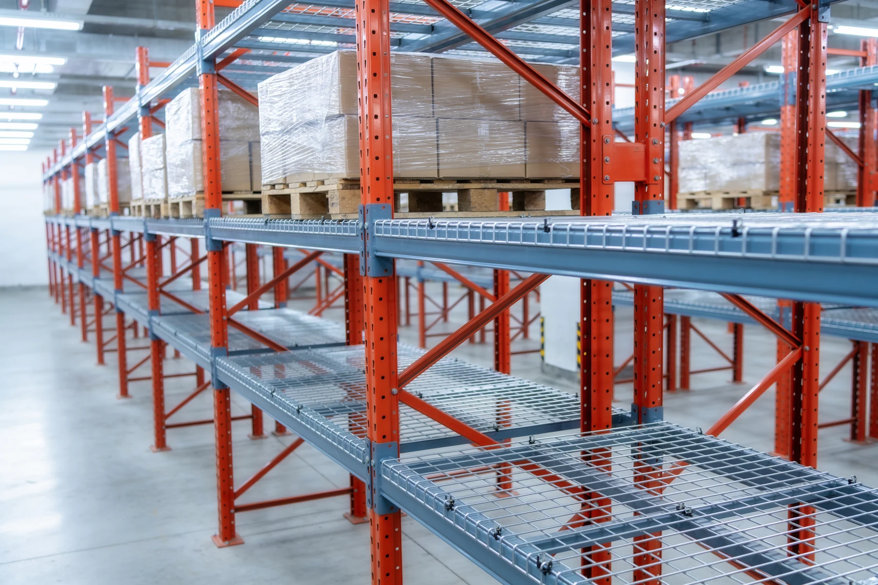 Bay of selective pallet racking with galvanized welded wire mesh decking laid across the beam levels, pallets of stretch-wrapped cartons stored on the upper decks